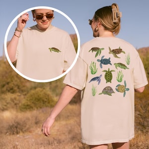May include: Cream-colored t-shirt featuring a small turtle graphic on the front and a larger design of various turtles and seaweed on the back. The turtles are in shades of green, brown, and blue. The shirt is a relaxed fit.