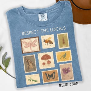 May include: Blue jean-colored t-shirt with the text "RESPECT THE LOCALS." The shirt features a collection of postage stamp designs with illustrations of insects, plants, and animals. The shirt is a Comfort Colors brand.