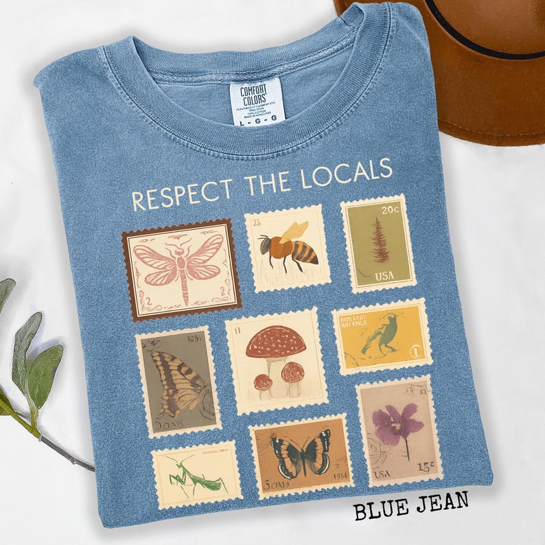 May include: Blue jean-colored t-shirt with the text "RESPECT THE LOCALS." The shirt features a collection of postage stamp designs with illustrations of insects, plants, and animals. The shirt is a Comfort Colors brand.