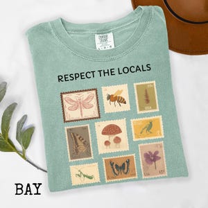 May include: A light green t-shirt with the text "RESPECT THE LOCALS" and various postage stamp designs featuring insects, plants, and a bird. The shirt is folded and has a Comfort Colors tag.