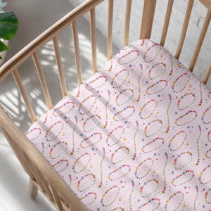 Puede incluir: Una sábana rosa claro para cuna con un patrón repetido de óvalos coloridos y líneas punteadas. La sábana está dentro de una cuna de madera con un acabado de madera clara. Una planta verde en una maceta blanca es visible en el fondo.