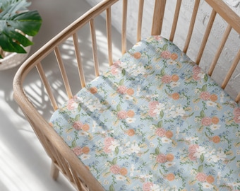 Peut inclure: Un drap de lit pour bébé bleu clair avec un motif floral. Le motif présente des roses roses et oranges, des fleurs blanches et des accents bleus. Le drap est dans un lit bébé en bois clair.