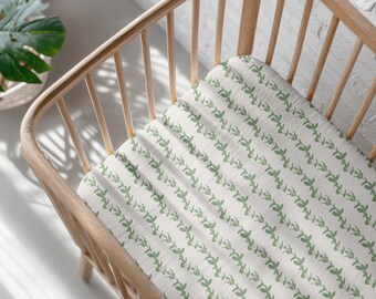 Peut inclure: Un drap-housse pour lit bébé avec un motif répété de feuilles vertes sur fond blanc. Le drap est dans un lit bébé en bois clair. Une plante verte dans un pot blanc est visible en arrière-plan.