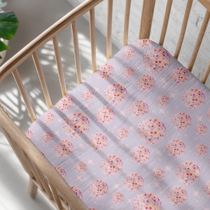 May include: A light purple fitted crib sheet with a repeating pattern of disco ball-like shapes in pink, yellow, and red. The sheet also features small star shapes. The sheet is inside a light wooden crib.