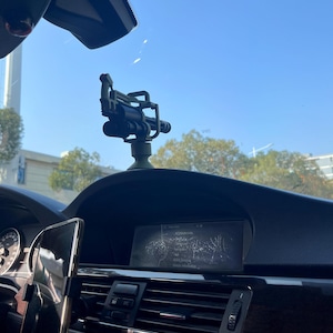Pu&ograve; includere: Cruscotto nero di un'auto con supporto per telefono e un piccolo oggetto decorativo verde scuro e nero che assomiglia a una miniatura di un'arma. L'interno include uno schermo di navigazione e bocchette dell'aria. Il parabrezza e il cielo blu sono visibili.