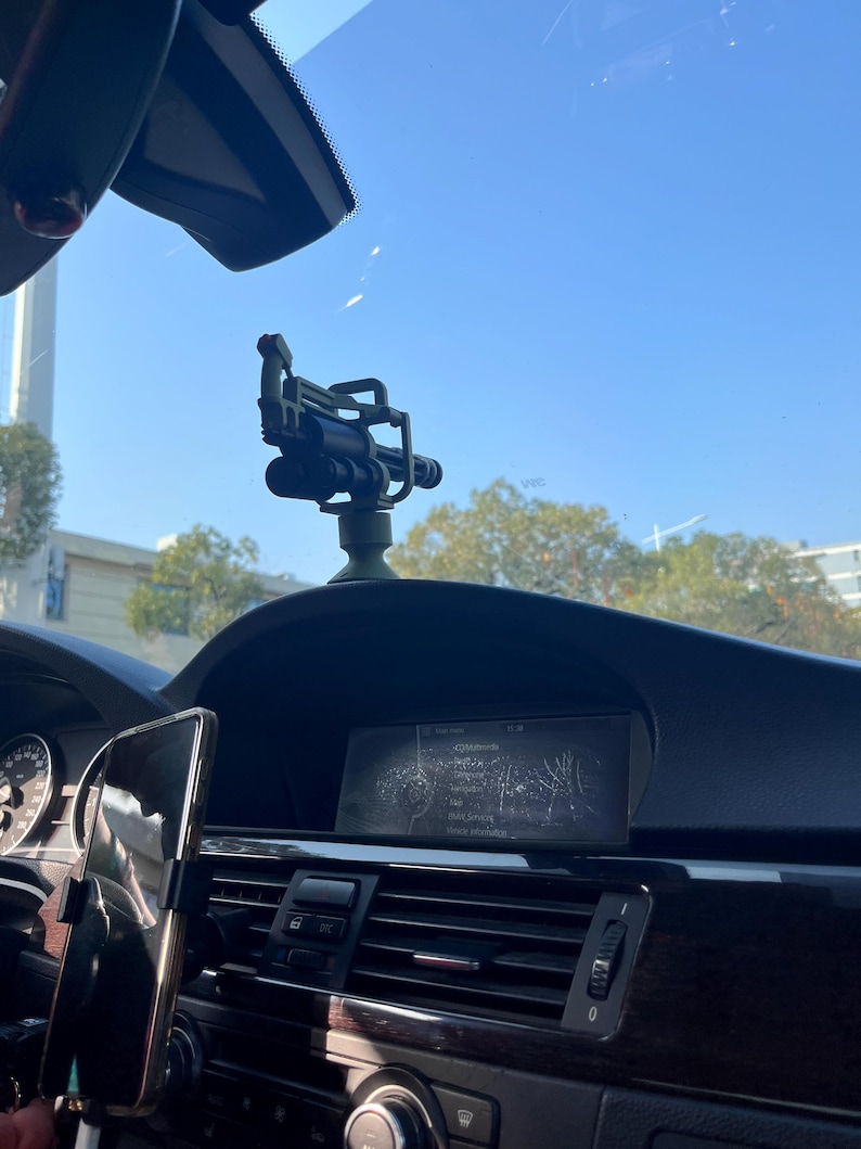 Pu&ograve; includere: Cruscotto nero di un'auto con supporto per telefono e un piccolo oggetto decorativo verde scuro e nero che assomiglia a una miniatura di un'arma. L'interno include uno schermo di navigazione e bocchette dell'aria. Il parabrezza e il cielo blu sono visibili.
