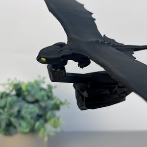 May include: A black dragon figurine with outstretched wings and yellow eyes. The dragon is mounted on a black base. The figurine is a detailed representation of a dragon, with textured wings and a sleek body. The dragon is a fantasy-themed item.