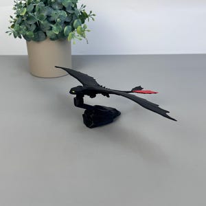 May include: A black dragon figurine with outstretched wings and a red tail fin. The dragon is perched on a dark base. A small green plant in a beige pot is in the background.