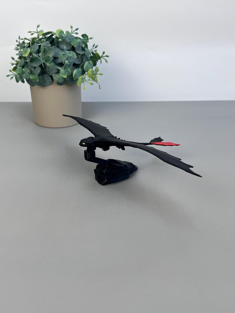 May include: A black dragon figurine with outstretched wings and a red tail fin. The dragon is perched on a dark base. A small green plant in a beige pot is in the background.