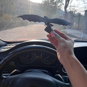 May include: A black dragon figurine with outstretched wings is held up against a bright, sunny background. The dragon is perched on a small base. The image is taken from inside a car, with the steering wheel and dashboard visible.