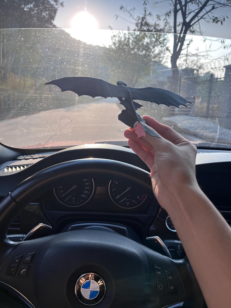 May include: A black dragon figurine with outstretched wings is held up against a bright, sunny background. The dragon is perched on a small base. The image is taken from inside a car, with the steering wheel and dashboard visible.