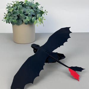 May include: A black dragon figurine with outstretched wings and a red tail fin. The dragon is positioned in front of a small potted plant with green foliage. The figurine has a sleek, stylized design.