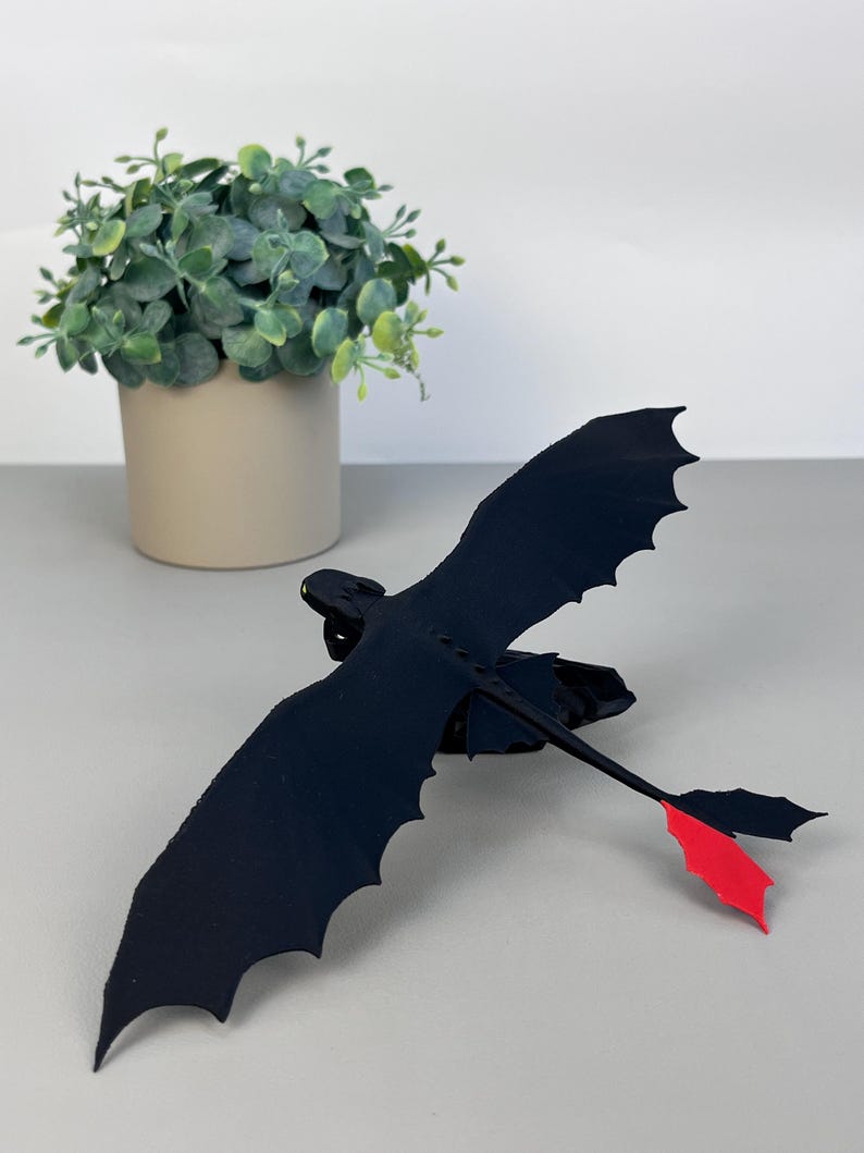 May include: A black dragon figurine with outstretched wings and a red tail fin. The dragon is positioned in front of a small potted plant with green foliage. The figurine has a sleek, stylized design.