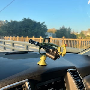 May include: A miniature, olive green and black Gatling gun model, mounted on a yellow-green base. The model is detailed with a rotating barrel and a cylindrical magazine. It is positioned on a car dashboard.