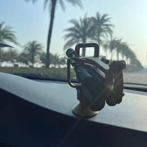 Pu&ograve; includere: Un supporto per telefono color oro con base a ventosa, progettato per essere fissato al cruscotto di un'auto. Il supporto ha un braccio flessibile e un alloggiamento per fissare il telefono. Palme visibili sullo sfondo.