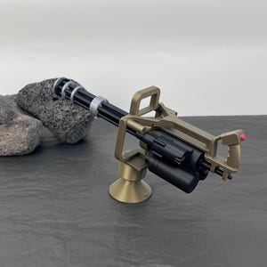 Pu&ograve; includere: Un modello in miniatura di mitragliatrice Gatling color bronzo con canna nera e caricatore cilindrico. L'arma &egrave; montata su una base di bronzo e presenta un'impugnatura con un pulsante rosso. Il modello &egrave; ambientato su uno sfondo neutro.