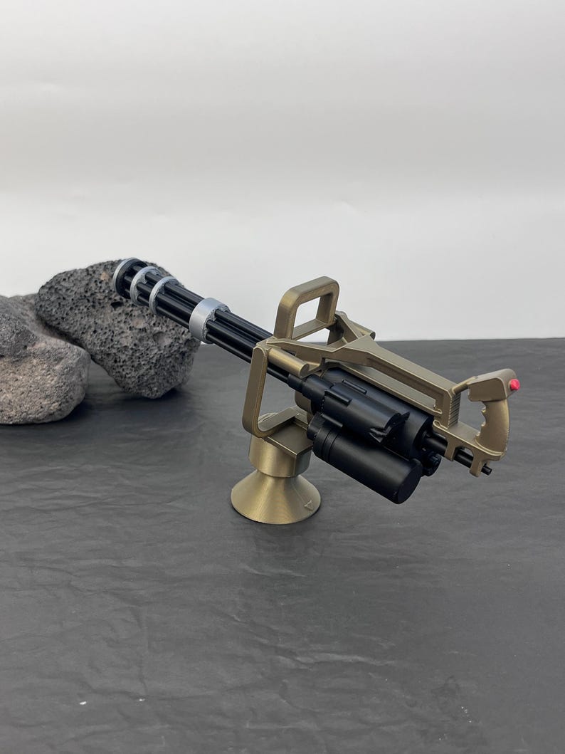 Pu&ograve; includere: Un modello in miniatura di mitragliatrice Gatling color bronzo con canna nera e caricatore cilindrico. L'arma &egrave; montata su una base di bronzo e presenta un'impugnatura con un pulsante rosso. Il modello &egrave; ambientato su uno sfondo neutro.
