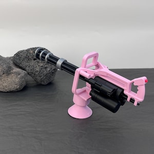 Pu&ograve; includere: Una minigun giocattolo rosa e nera. La pistola presenta un telaio rosa, una canna nera con dettagli argentati e un tamburo di munizioni nero. Il giocattolo &egrave; posto su una superficie scura con uno sfondo chiaro.