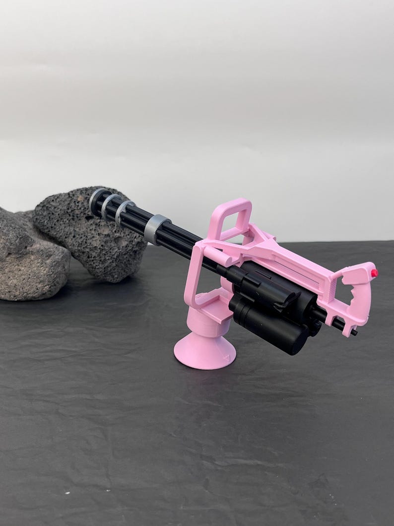 Pu&ograve; includere: Una minigun giocattolo rosa e nera. La pistola presenta un telaio rosa, una canna nera con dettagli argentati e un tamburo di munizioni nero. Il giocattolo &egrave; posto su una superficie scura con uno sfondo chiaro.