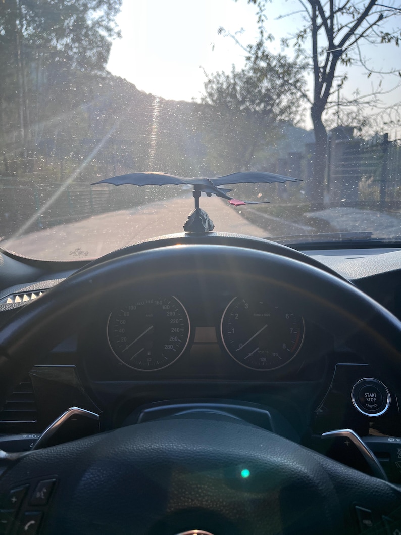 May include: A black dragon car ornament with outstretched wings, mounted on a dashboard. The dragon has a red accent on its tail. The car's steering wheel and dashboard gauges are visible in the foreground, with a road and trees in the background.