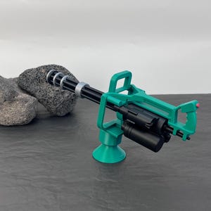 Pu&ograve; includere: Una minigun giocattolo verde e nera con canna rotante e maniglia. La pistola &egrave; montata su una base verde. Lo sfondo &egrave; una superficie grigio scuro con uno sfondo grigio chiaro.