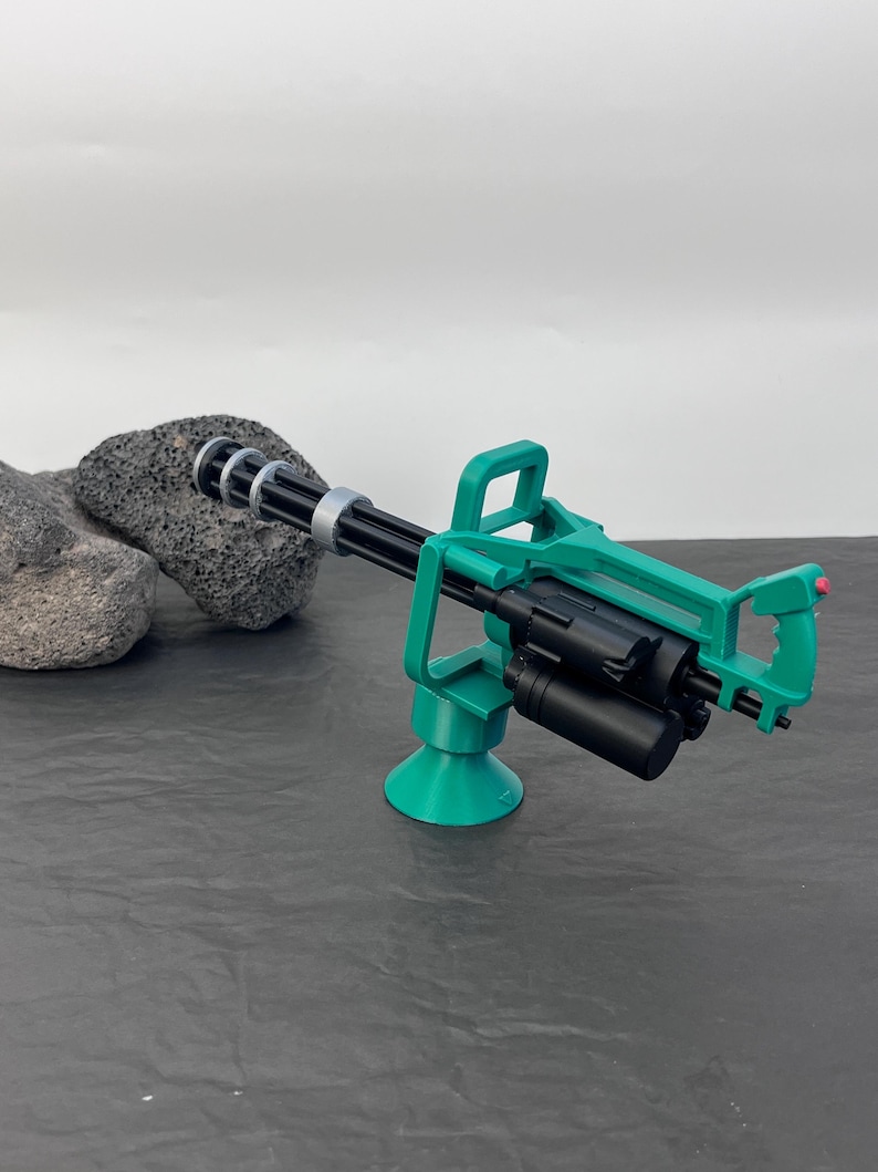 Pu&ograve; includere: Una minigun giocattolo verde e nera con canna rotante e maniglia. La pistola &egrave; montata su una base verde. Lo sfondo &egrave; una superficie grigio scuro con uno sfondo grigio chiaro.