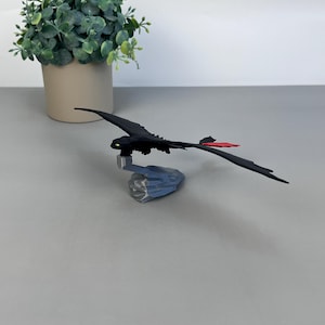 May include: A black dragon figurine with a red tail fin and green eyes. The dragon is perched on a gray, rocky base. A small, green plant in a beige pot is in the background.