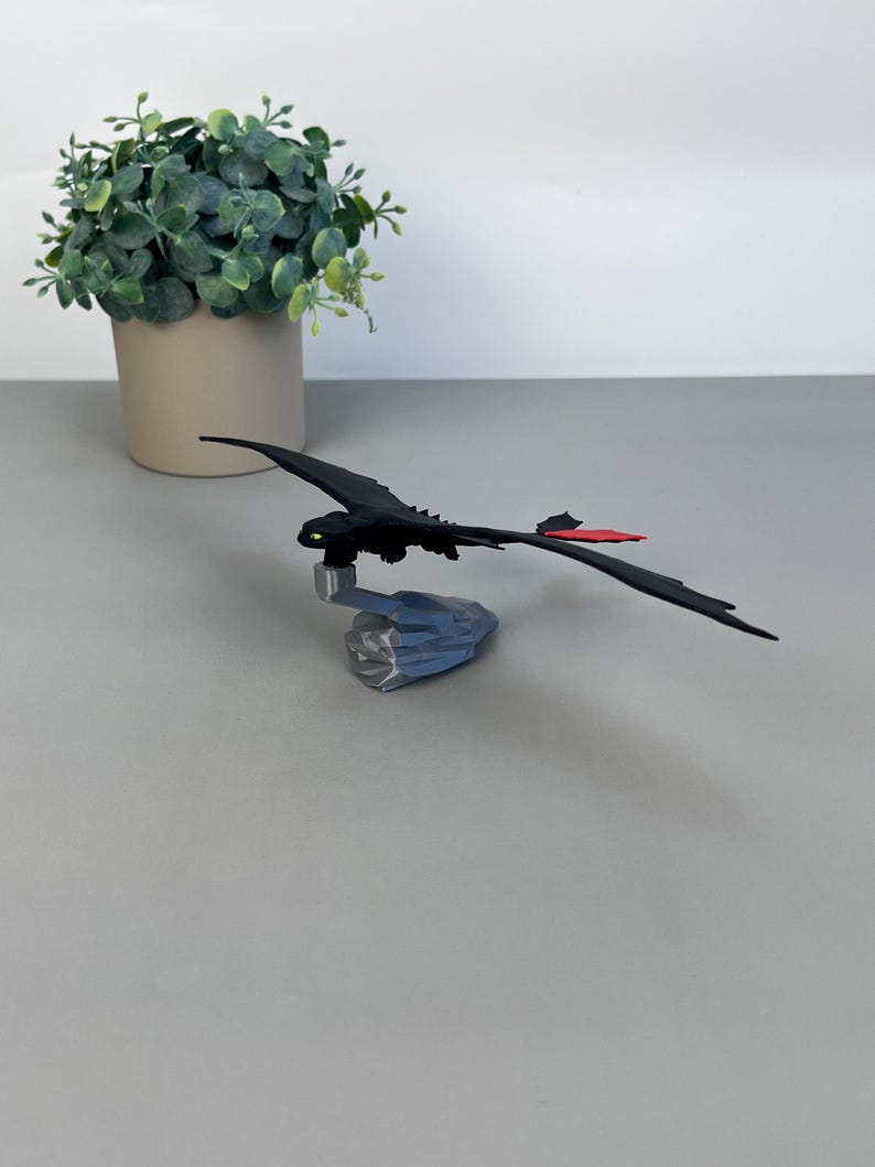 May include: A black dragon figurine with a red tail fin and green eyes. The dragon is perched on a gray, rocky base. A small, green plant in a beige pot is in the background.
