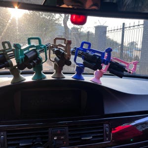 Pu&ograve; includere: Una collezione di mitragliatrici giocattolo in miniatura in vari colori, tra cui verde, verde acqua, marrone, blu e rosa. I giocattoli sono montati su supporti ed esposti sul cruscotto di un'auto. L'immagine &egrave; stata scattata alla luce del giorno.