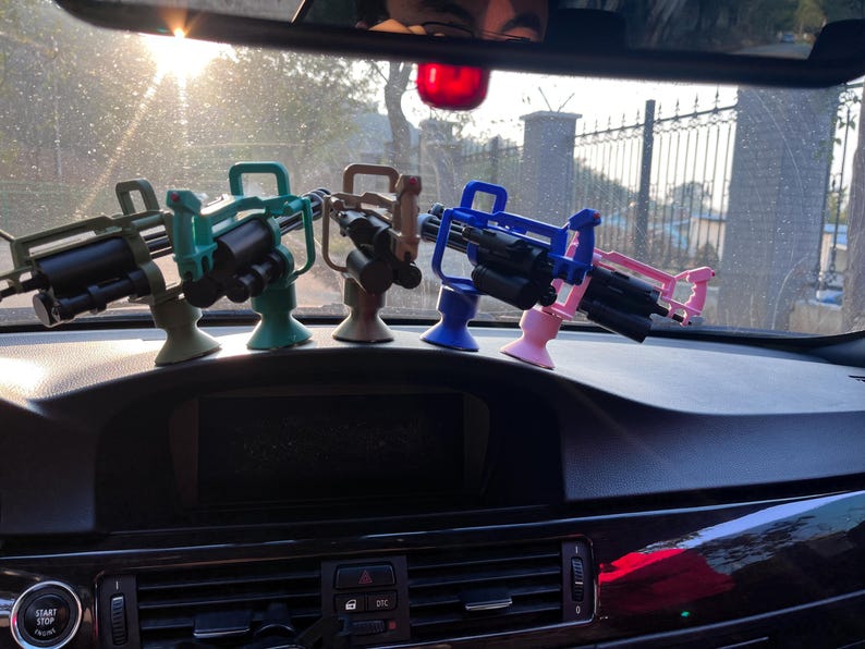 Pu&ograve; includere: Una collezione di mitragliatrici giocattolo in miniatura in vari colori, tra cui verde, verde acqua, marrone, blu e rosa. I giocattoli sono montati su supporti ed esposti sul cruscotto di un'auto. L'immagine &egrave; stata scattata alla luce del giorno.