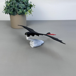 May include: A black dragon figurine with a red tail fin and green eyes, perched on a white stone base. The dragon is set against a neutral gray background, with a potted green plant in the background.