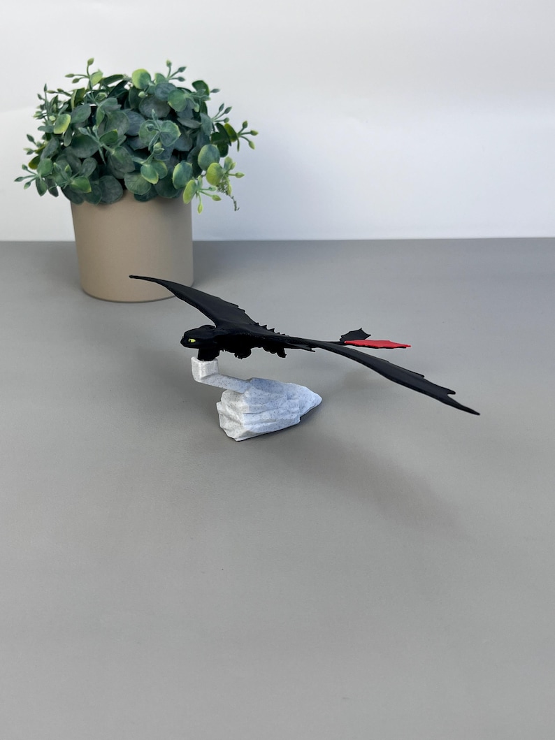 May include: A black dragon figurine with a red tail fin and green eyes, perched on a white stone base. The dragon is set against a neutral gray background, with a potted green plant in the background.