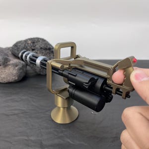 Pu&ograve; includere: Un modello in miniatura di mitragliatrice Gatling color oro con canna e impugnatura nere. Il modello ha una base cilindrica e un'impugnatura con un pulsante rosso. La pistola &egrave; tenuta in mano.