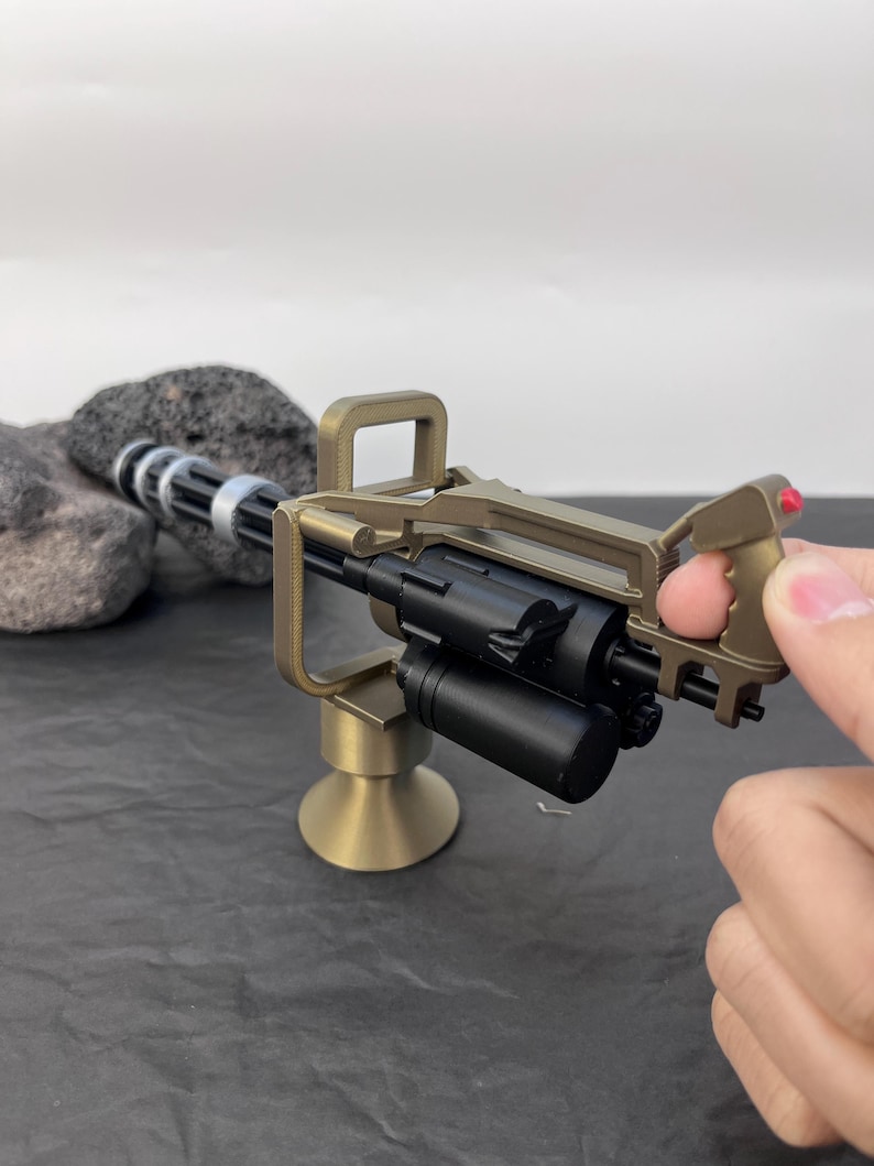 Pu&ograve; includere: Un modello in miniatura di mitragliatrice Gatling color oro con canna e impugnatura nere. Il modello ha una base cilindrica e un'impugnatura con un pulsante rosso. La pistola &egrave; tenuta in mano.