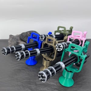 Pu&ograve; includere: Una collezione di mini-mitragliatrici Gatling in vari colori: blu, beige, verde, rosa e verde. Ogni arma ha pi&ugrave; canne, una canna nera e una base circolare. Le armi sono esposte su una superficie scura.