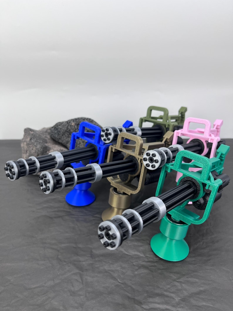 Pu&ograve; includere: Una collezione di mini-mitragliatrici Gatling in vari colori: blu, beige, verde, rosa e verde. Ogni arma ha pi&ugrave; canne, una canna nera e una base circolare. Le armi sono esposte su una superficie scura.