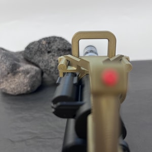Pu&ograve; includere: Primo piano di un'arma da fuoco color cachi e nera con mirino a punto rosso. L'arma da fuoco &egrave; posta su uno sfondo grigio con due rocce grigio scuro. L'attenzione &egrave; focalizzata sul mirino anteriore e sulla canna dell'arma.
