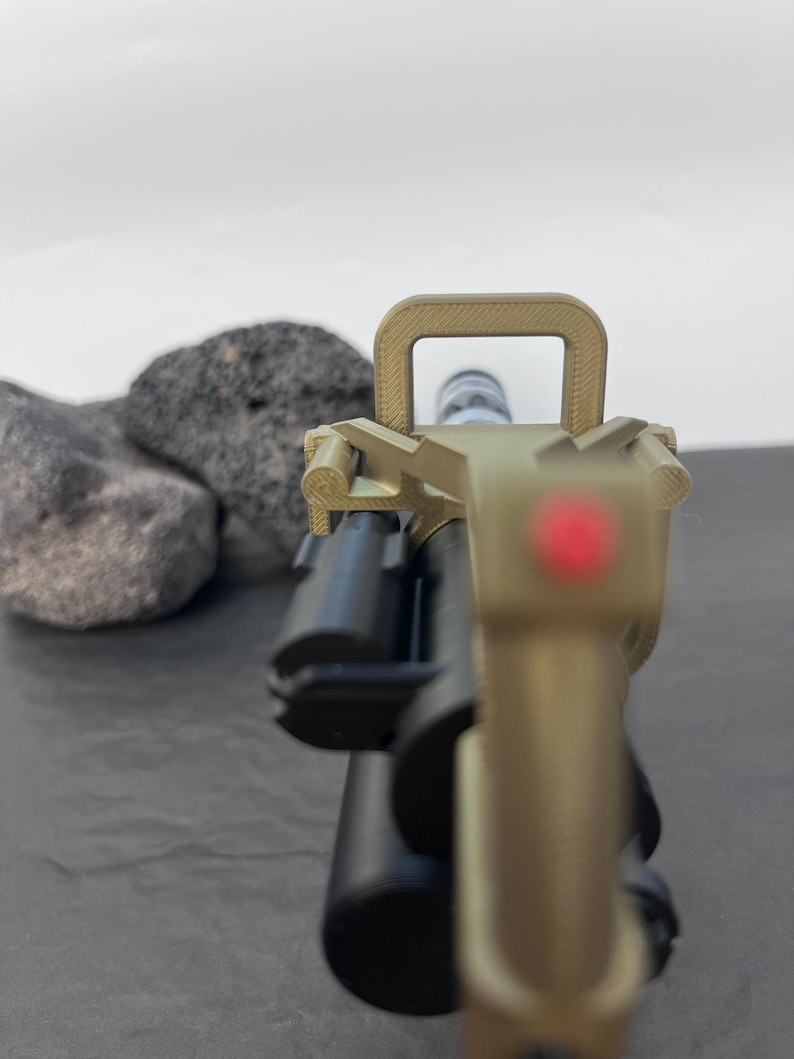 Pu&ograve; includere: Primo piano di un'arma da fuoco color cachi e nera con mirino a punto rosso. L'arma da fuoco &egrave; posta su uno sfondo grigio con due rocce grigio scuro. L'attenzione &egrave; focalizzata sul mirino anteriore e sulla canna dell'arma.
