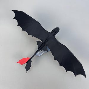 May include: A black dragon toy with outstretched wings and a red tail fin. The dragon is made of a soft material and has a detailed design. The wingspan is approximately 12 inches.