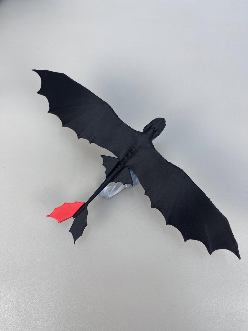 May include: A black dragon toy with outstretched wings and a red tail fin. The dragon is made of a soft material and has a detailed design. The wingspan is approximately 12 inches.