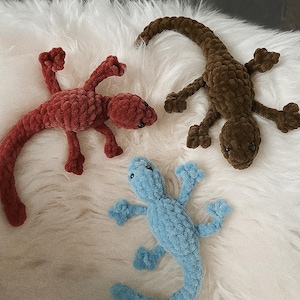 Peut inclure: Trois lézards en crochet faits à la main, de couleurs rouge, marron et bleu. Les lézards ont des pattes, des queues et des têtes détaillées, disposés sur une surface blanche et duveteuse. Sculptures douces pour la décoration intérieure.