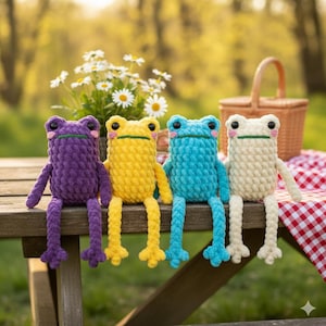 Könnte beinhalten: Vier handgefertigte Häkelfrosch-Spielzeuge in Lila, Gelb, Blau und Creme. Jeder Frosch hat ein lächelndes Gesicht und lange Beine und sitzt auf einer Holzoberfläche. Gänseblümchen und ein Picknickkorb sind im Hintergrund.