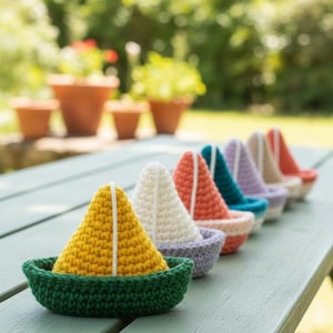 May include: A row of colorful, crocheted sailboats. Each boat features a different colored sail, including yellow, white, orange, teal, purple, and beige. The boats sit in a green, gray, and beige crocheted boat base. The sailboats are displayed on a weathered, light green picnic table.