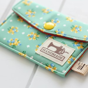 Może przedstawiać: Ręcznie robiony, miętowy portfel z tkaniny z kwiatowym wzorem z żółtych i niebieskich kwiatów. Portfel ma czerwone wykończenie, żółte zapięcie na guzik i etykietę "HANDMADE" z grafiką maszyny do szycia. Dołączona jest mała miarka.