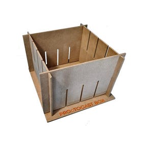Puede incluir: Una caja de madera ensamblada de color marrón claro con ranuras verticales cortadas en los lados. La caja está construida con varias piezas entrelazadas. El texto "PROCTOCAST BOX" está impreso en naranja en la base.