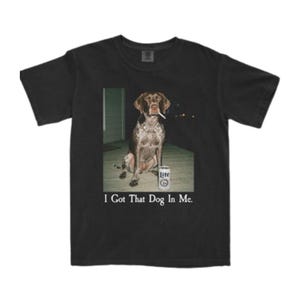 Op de afbeelding: Een zwart t-shirt met korte mouwen en een grafische print van een bruin-witte gevlekte hond die op een veranda zit met een sigaret in de bek en een blikje Lite-bier ernaast. De tekst "I Got That Dog In Me." staat onder de afbeelding gedrukt.