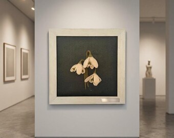 May include: A square framed artwork featuring three white snowdrop flowers with green stems centered on a dark charcoal background. The frame is a light-colored wood with a visible grain and a small rectangular metal plaque at the bottom right corner.