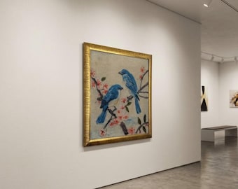 Puede incluir: Una pintura cuadrada en un marco dorado texturizado que representa dos pájaros azules posados en ramas con pequeñas flores rosas. La obra de arte se exhibe en una pared blanca lisa dentro de una galería, con un fondo claro y pinceladas visibles.