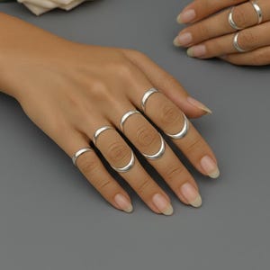 May include: Silver rings in various styles adorn a hand, showcasing a collection of jewelry. The rings include simple bands and curved designs, reflecting light against a gray backdrop. The image is a close-up, highlighting the details of the rings.