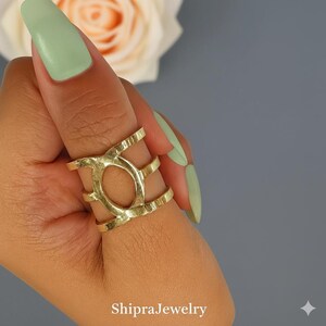 May include: A gold-toned ring featuring a wide, multi-band design with a central oval cutout. The metal surface has a textured, hammered finish. The ring spans across three finger joints, creating a bold, open-work appearance on the finger.